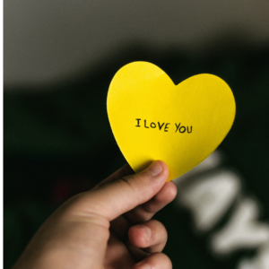 Käsi pitelee keltaista paperisydäntä, jossa lukee i love you.